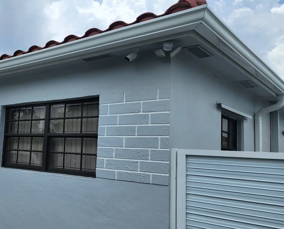Security cameras under the eave of a grey stucco home with white gutters