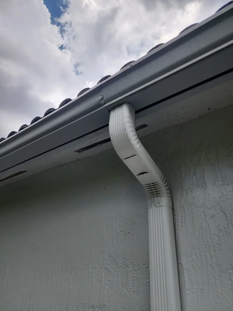 A white downspout elbow under a dark gutter on grey stucco