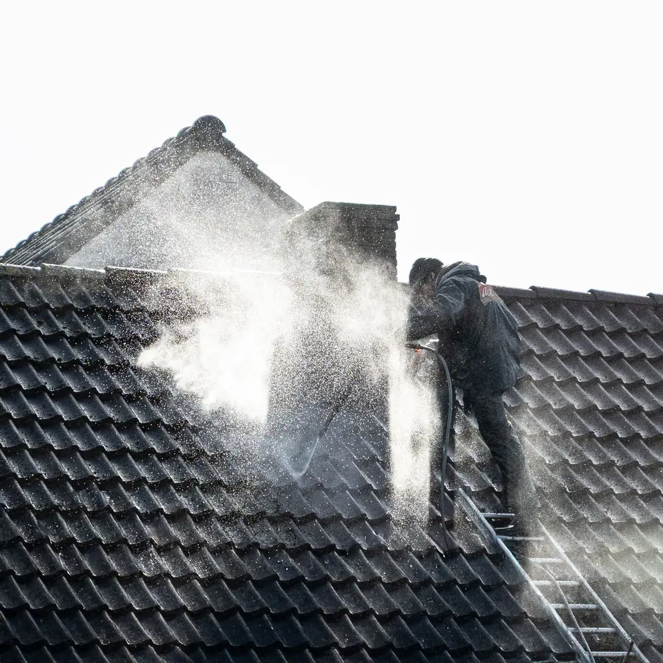 A worker washing a dark concrete tile roof from a ladder